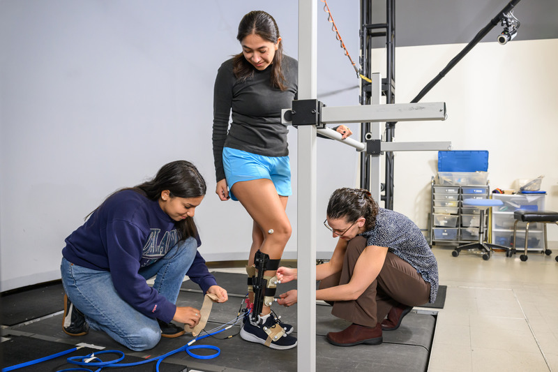 Elisa Arch (right), associate professor of kinesiology and applied physiology, and biomechanics and movement science doctoral student Zahra McKee outfit Iris Sanchez, a senior mechanical engineering major, with an ankle-foot-orthosis (AFO) equipped with sensors to monitor tendon load as part of a novel approach to help those with Achilles tendinopathy recover faster.