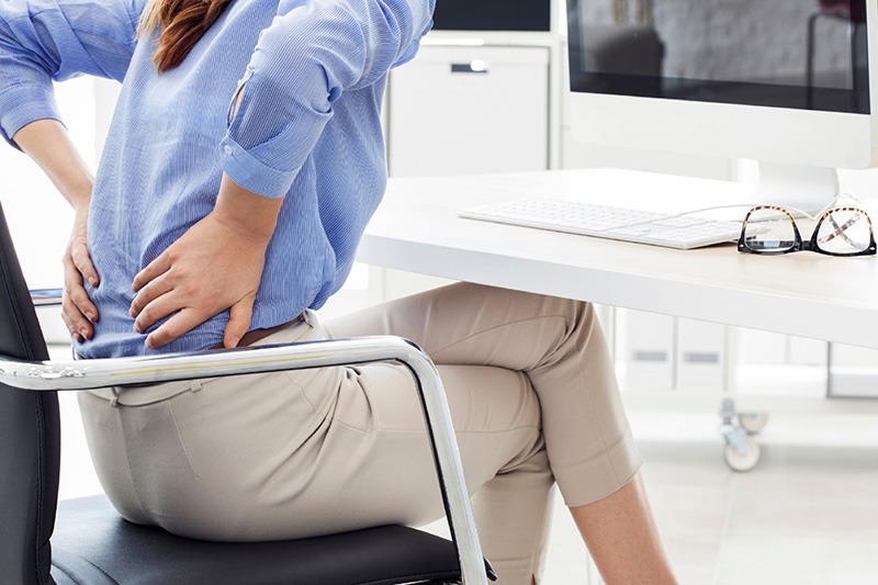 A woman seated at a desk in front of a computer holds her lower back with both hands. Source: https://pixabay.com/photos/back-pain-body-pain-ergonomics-6949392/